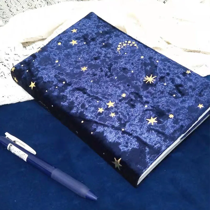 Couvre-livre bleu nuit au toucher velouté parsemé d'étoiles dorées, posé près d'un stylo