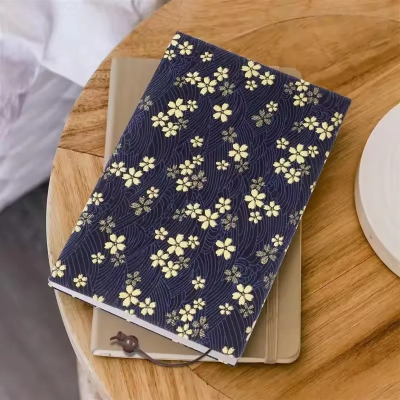Couvre-livre en tissu bleu nuit à fleurs de cerisier crème posé sur un carnet, sur une table en bois